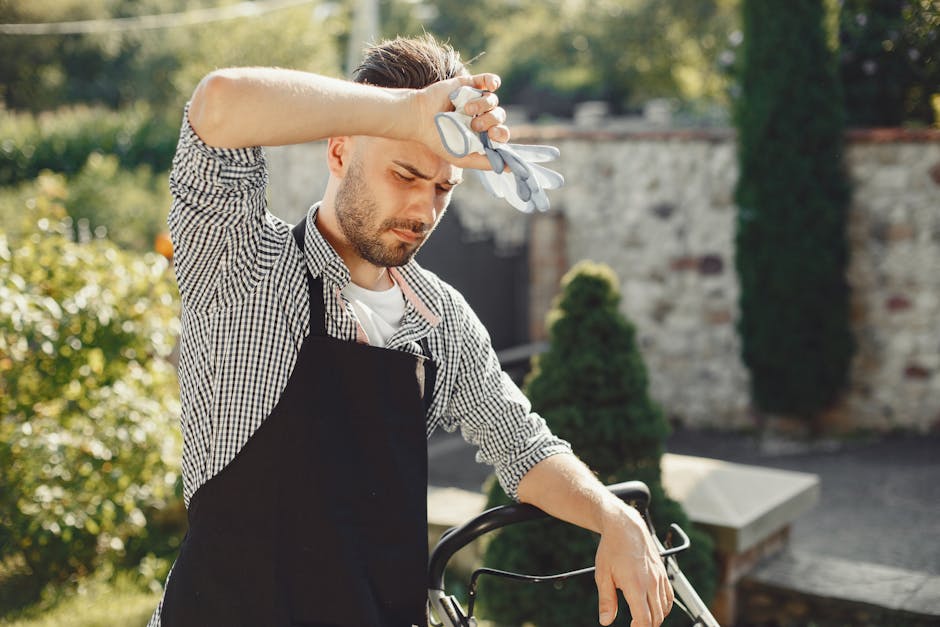 A man standing outdoors in a garden during daytime, holding his forehead with one hand and wearing gardening gloves while looking down with a focused expression. He is dressed in a black apron over a 
