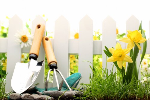 A close-up view of a small, neatly maintained garden section featuring a white picket fence in the background, with vibrant yellow daffodils and white daisies growing along the base. In the foreground, gardening tools including a hand trowel, a hand fork, and a pair of gardening gloves are resting on the soil, which is dark and well-turned, indicating recent planting or cultivation activity. The grass nearby is lush, green, and slightly overgrown, contrasting with the bright flowers and clean fence. Natural daylight illuminates the scene, suggesting a sunny day, and the overall setting reflects an organized outdoor space suitable for garden maintenance or lawn care services offered by Lawn Mowing Charlton in the Charlton area, enhancing the natural beauty of a typical UK garden environment.